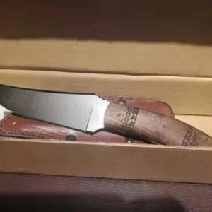 May include: A hunting knife with a polished steel blade and a wooden handle with decorative carvings. The knife rests in a brown leather sheath inside a cardboard box.