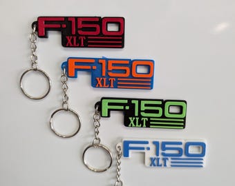 Puede incluir: Cuatro llaveros con el texto "F-150 XLT" en rojo, naranja, verde y azul. Cada llavero tiene una cadena plateada y un llavero. Los llaveros están sobre un fondo blanco.