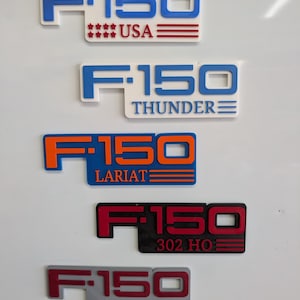 Può includere: Una collezione di emblemi F-150 in vari colori. Gli emblemi includono "USA", "Thunder", "Lariat", "302 HO" e "Dwight". Ogni emblema presenta il logo "F-150".