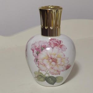 May include: A white ceramic fragrance lamp with a rounded body featuring a botanical illustration of pink and yellow roses with green leaves. The lamp is topped with a polished gold-colored metal crown cap with vertical cutouts for ventilation.