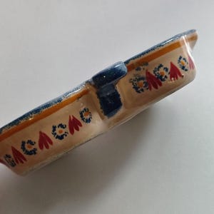 May include: A rectangular ceramic dish with a divided interior. The dish features a beige base with a repeating pattern of red and blue floral designs. The rim is accented with blue and orange stripes. A blue handle is on the side.