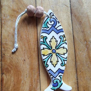 May include: A ceramic fish-shaped ornament with a decorative floral design in yellow, blue, and teal on a white background. The ornament has a string and two wooden beads attached for hanging. The fish is approximately 5 inches long.