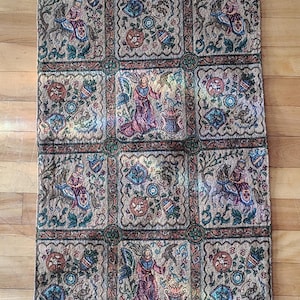 May include: A long, rectangular woven wall hanging featuring a repeating grid pattern of medieval-inspired scenes. The tapestry displays figures on horseback, heraldic shields, and standing figures with birds, rendered in muted earth tones.