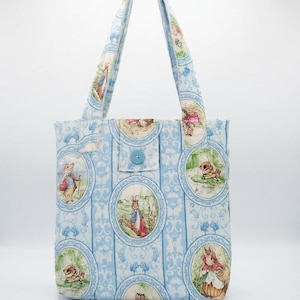 May include: A light blue fabric tote bag featuring a repeating pattern of oval frames containing illustrations of rabbits in various outdoor settings. The bag has two matching fabric handles and a central flap closure secured with a single blue button.