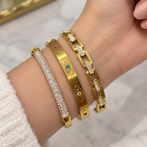 May include: A set of three gold-toned bracelets. One is a solid band covered in clear crystals, the second is a flat band featuring engraved eye motifs with small coloured stones, and the third is a chain-link style bracelet with crystal accents.