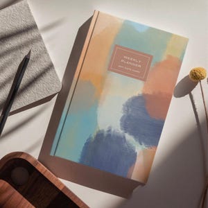 Könnte beinhalten: Ein Hardcover-Wochenplaner mit einem abstrakten Aquarelldesign in Orange-, Blau- und Grüntönen. Der Einband trägt den Text "WEEKLY PLANNER ANY DATE DIARY" in einem rechteckigen Rahmen. Ein Bleistift und ein Notizblock sind sichtbar.