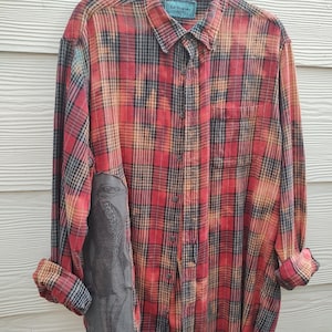 May include: A long-sleeve button-down flannel shirt in a red, black, and yellow plaid pattern with a bleached effect. The shirt features a dark gray rectangular patch on the lower right side displaying a detailed illustration of a standing penguin.