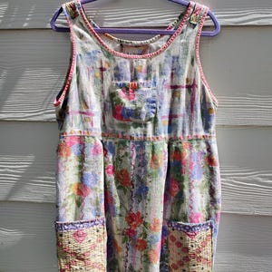May include: A sleeveless, floral-print dress with a scoop neckline and two front pockets. The dress features a patchwork design with pink, blue, and yellow floral patterns. The dress is hanging on a purple hanger.