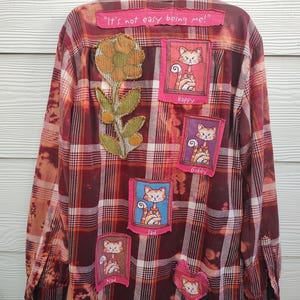 May include: A long-sleeve button-down flannel shirt in maroon and white plaid with a bleached effect. The back features a fabric flower appliqué, a heart patch, and four rectangular patches of cats labeled Happy, Giddy, Sad, and Mad, with the text "It's not easy being me!" at the top.