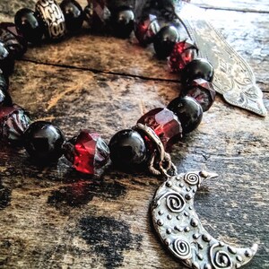 May include: A beaded bracelet featuring alternating round black beads, faceted translucent red beads, and a silver-toned metal spacer bead. A silver-toned crescent moon charm with a textured spiral pattern hangs from the bracelet, alongside a flat metal charm.