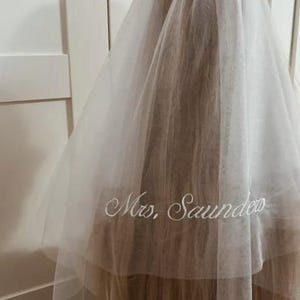 May include: A sheer white bridal veil made of fine tulle fabric, draped over long hair. The lower section of the veil features the embroidered text "Mrs. Saunders" in a cursive white script font, positioned horizontally across the center.