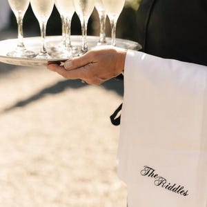May include: A silver serving tray holding five clear glass champagne flutes filled with a sparkling beverage. A white cloth napkin with the embroidered black text The Riddles is draped over the arm of the server wearing a black uniform.