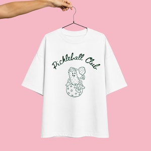 K&ouml;nnte beinhalten: Ein wei&szlig;es Kurzarm-T-Shirt mit einer dunkelgr&uuml;nen Grafik einer Comic-Gew&uuml;rzgurke, die eine Kappe und Turnschuhe tr&auml;gt und einen Pickleball-Schl&auml;ger h&auml;lt, w&auml;hrend sie auf einem Ball sitzt. Der Text "Pickleball Club" ist in Dunkelgr&uuml;n &uuml;ber der Grafik gedruckt.