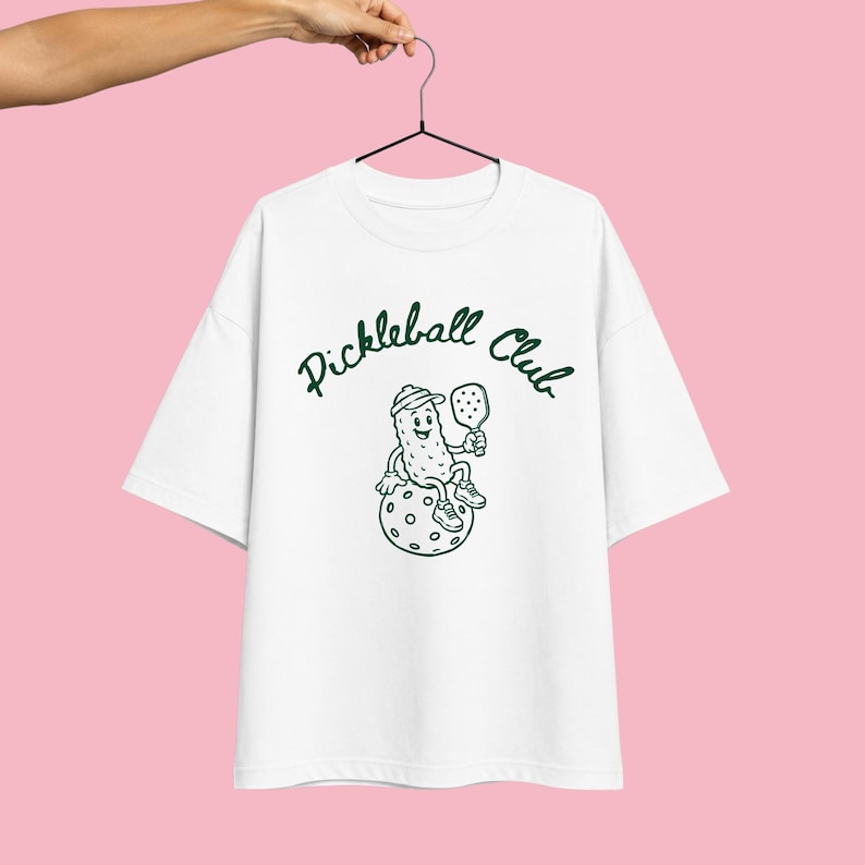 K&ouml;nnte beinhalten: Ein wei&szlig;es Kurzarm-T-Shirt mit einer dunkelgr&uuml;nen Grafik einer Comic-Gew&uuml;rzgurke, die eine Kappe und Turnschuhe tr&auml;gt und einen Pickleball-Schl&auml;ger h&auml;lt, w&auml;hrend sie auf einem Ball sitzt. Der Text "Pickleball Club" ist in Dunkelgr&uuml;n &uuml;ber der Grafik gedruckt.