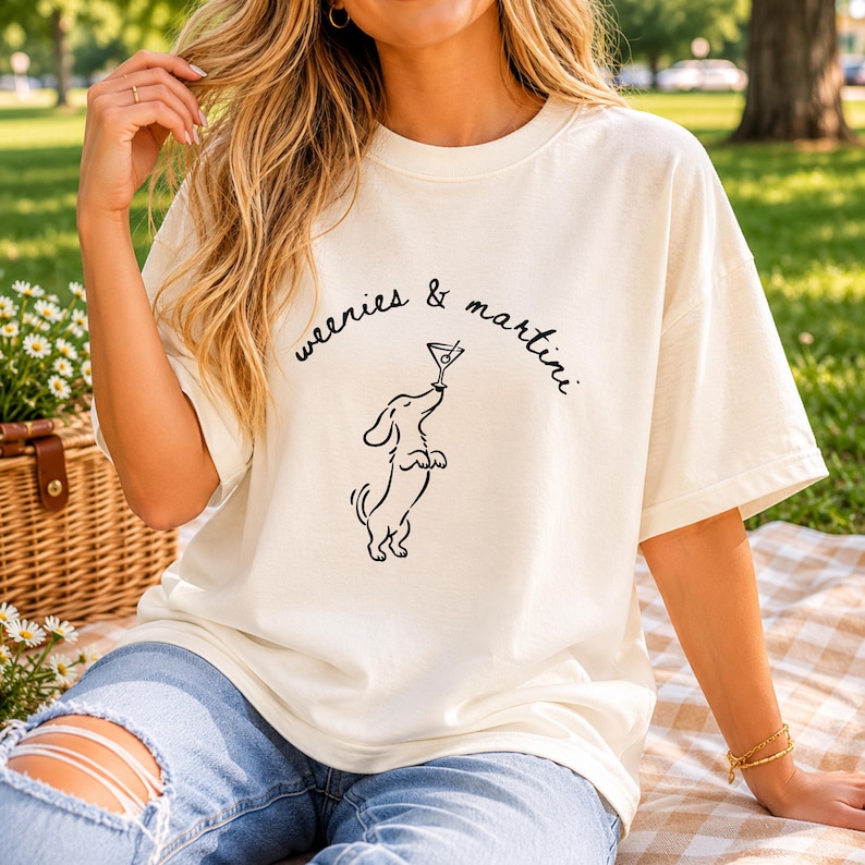 May include: An off-white short-sleeve t-shirt featuring a black line drawing of a dachshund standing on its hind legs while balancing a martini glass on its nose, with the text "weenies & martini" printed in a black cursive script above.