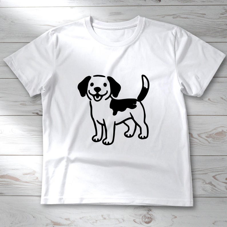 Op de afbeelding: Wit T-shirt met een zwart-witte cartoonhondenillustratie. De hond heeft zwarte oren, een zwarte vlek op zijn rug en een lachende uitdrukking. Het T-shirt is gemaakt van een zacht materiaal en heeft korte mouwen.
