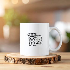 Op de afbeelding: Witte keramische mok met een zwarte lijntekening van een lachende bulldog. De mok heeft een gebogen handvat en staat op een ronde houten onderzetter. De achtergrond is vervaagd met natuurlijk licht.