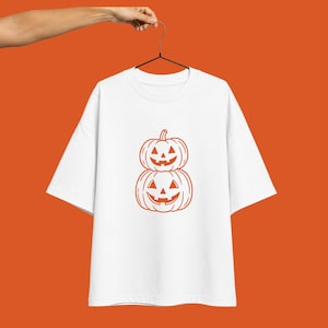 May include: A white short-sleeved t-shirt featuring a graphic print of two stacked orange jack-o'-lantern pumpkins. The design is centered on the chest of the shirt, which is displayed on a black hanger against a solid orange background.