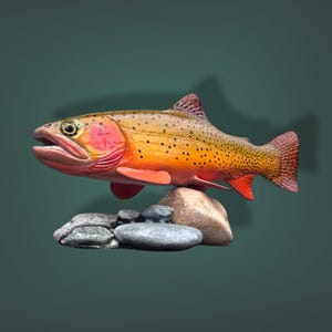 May include: A detailed sculpture of a trout fish with vibrant orange, yellow, and red hues, speckled with black dots. The fish rests on a collection of gray and tan stones, set against a dark green backdrop.