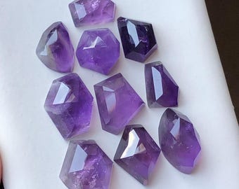 May include: A selection of faceted amethyst gemstones in various geometric shapes. The polished stones display a range of purple hues, from light lavender to deep violet. The gemstones are arranged on a white surface, showcasing their reflective surfaces.