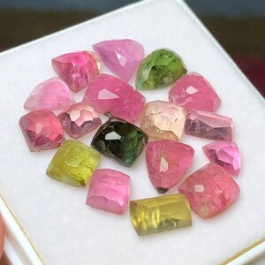 May include: A collection of faceted gemstones in various colors, including pink, green, and yellow, arranged on a white surface. The gemstones have a polished, crystalline appearance, with visible internal structures and light reflections. The assortment includes a variety of shapes and sizes.