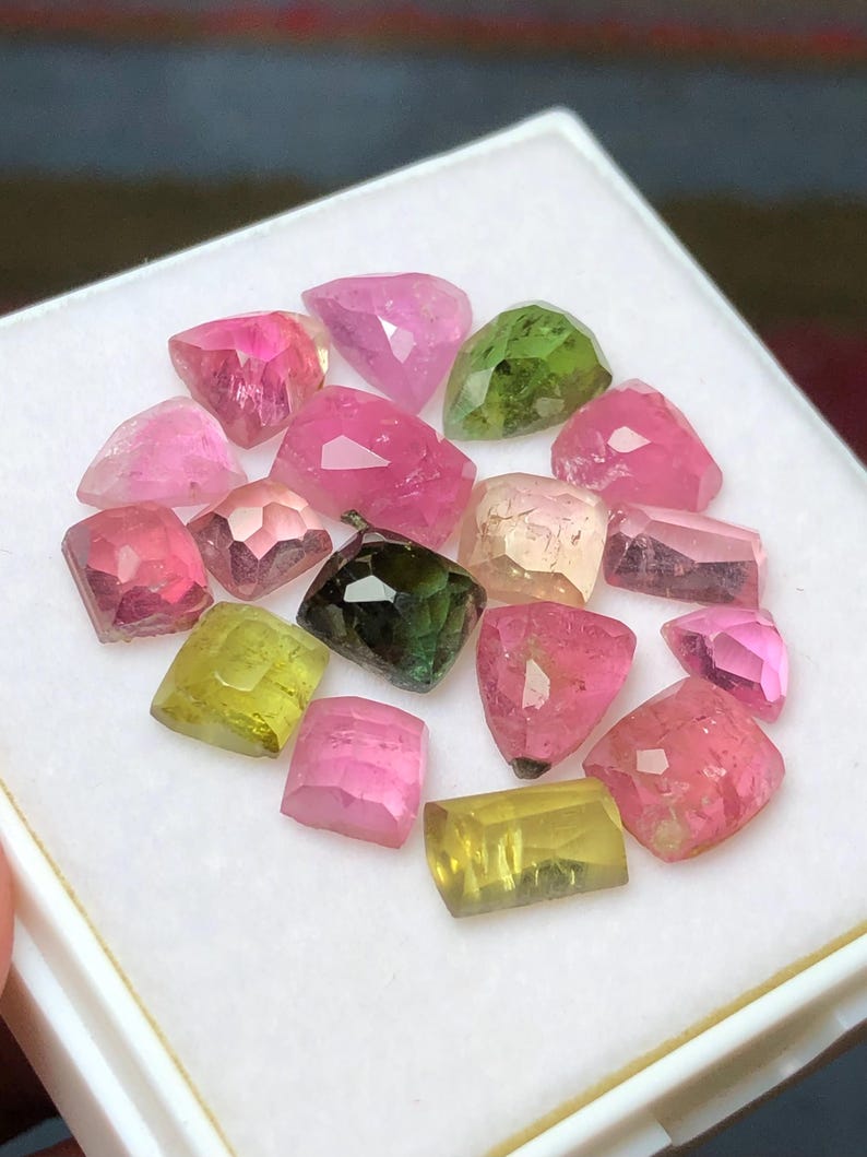 May include: A collection of faceted gemstones in various colors, including pink, green, and yellow, arranged on a white surface. The gemstones have a polished, crystalline appearance, with visible internal structures and light reflections. The assortment includes a variety of shapes and sizes.