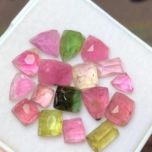May include: A collection of faceted tourmaline gemstones in various colors, including pink, green, and yellow. The gemstones are displayed in a white tray, showcasing their different shapes and sizes. The gems have a translucent quality.