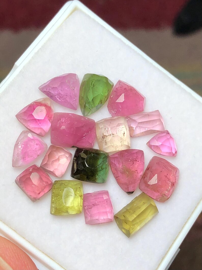 May include: A collection of faceted tourmaline gemstones in various colors, including pink, green, and yellow. The gemstones are displayed in a white tray, showcasing their different shapes and sizes. The gems have a translucent quality.
