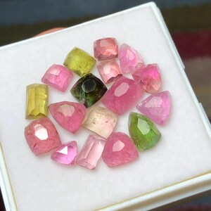 May include: A collection of faceted gemstones in various shades of pink, green, and yellow, displayed in a white box. The gemstones have different shapes, including rectangular, oval, and triangular cuts. The gemstones are arranged in a scattered pattern.