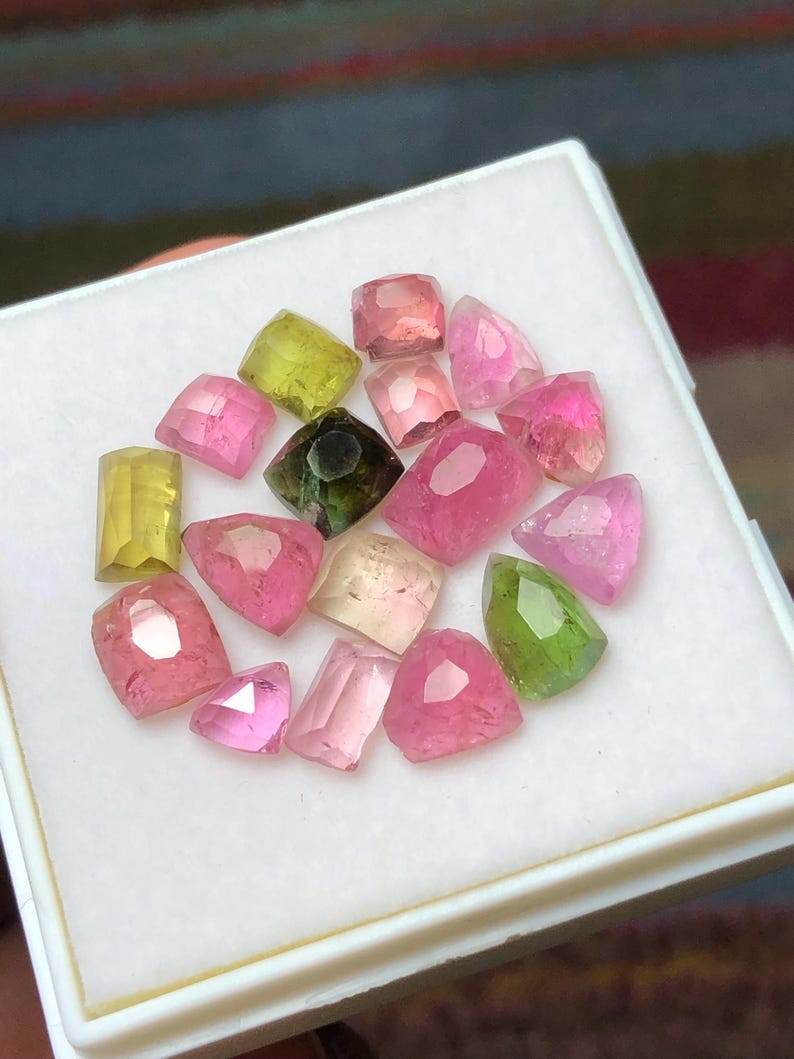 May include: A collection of faceted gemstones in various shades of pink, green, and yellow, displayed in a white box. The gemstones have different shapes, including rectangular, oval, and triangular cuts. The gemstones are arranged in a scattered pattern.