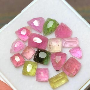 May include: A collection of faceted gemstones in various shapes and colors, including pink, green, and yellow. The gemstones are displayed in a white square container. The gems have a polished, reflective surface.