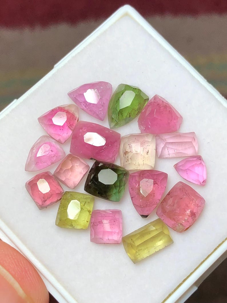 May include: A collection of faceted gemstones in various shapes and colors, including pink, green, and yellow. The gemstones are displayed in a white square container. The gems have a polished, reflective surface.