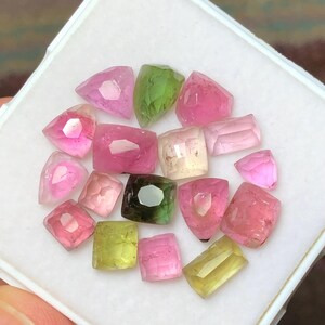 May include: A collection of faceted gemstones in various shapes and colors, including pink, green, and yellow. The gems are displayed in a white, square container, showcasing their clarity and brilliance. The gemstones are likely tourmaline.