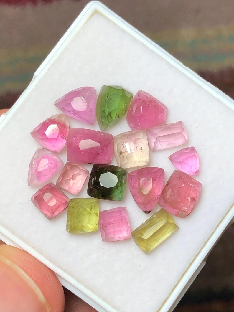 May include: A collection of faceted gemstones in various shapes and colors, including pink, green, and yellow. The gems are displayed in a white, square container, showcasing their clarity and brilliance. The gemstones are likely tourmaline.