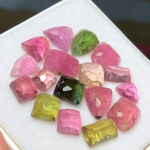 May include: A collection of faceted gemstones in various colors, including pink, green, and yellow. The gemstones are displayed on a white surface, showcasing their different shapes and sizes. The gems are likely tourmaline.