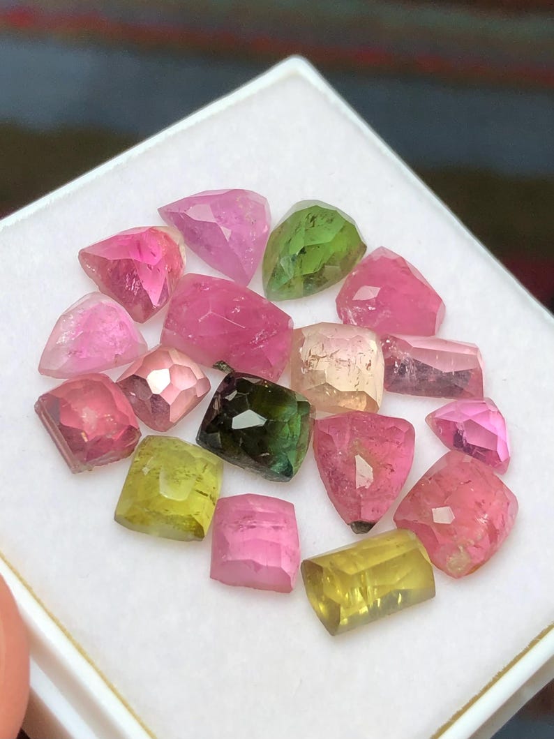 May include: A collection of faceted gemstones in various colors, including pink, green, and yellow. The gemstones are displayed on a white surface, showcasing their different shapes and sizes. The gems are likely tourmaline.