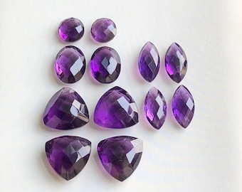 May include: A selection of faceted amethyst gemstones in various shapes, including oval, teardrop, and triangular cuts. The gemstones display a deep purple hue and are arranged on a white surface.