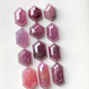 May include: A collection of faceted, hexagonal gemstones in varying shades of pink and red. The gemstones are arranged on a white surface, showcasing their polished surfaces and internal clarity. The gems have a geometric shape and are likely used for jewelry making.