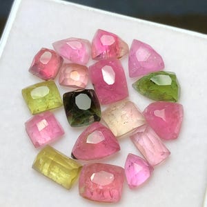 May include: A collection of faceted gemstones in various colors, including pink, green, yellow, and clear. The gemstones are arranged in a circular pattern within a white container. The gems have a polished, reflective surface.