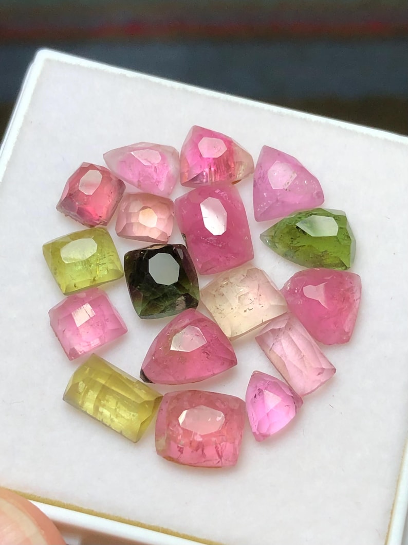 May include: A collection of faceted gemstones in various colors, including pink, green, yellow, and clear. The gemstones are arranged in a circular pattern within a white container. The gems have a polished, reflective surface.