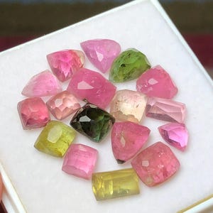 May include: A collection of faceted gemstones in various colors, including pink, green, and yellow, displayed in a white tray. The gemstones have a polished, reflective surface, and are of different shapes and sizes.