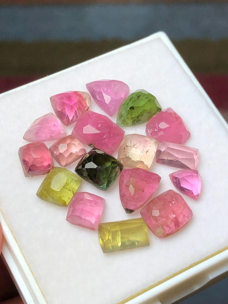 May include: A collection of faceted gemstones in various colors, including pink, green, and yellow, displayed in a white tray. The gemstones have a polished, reflective surface, and are of different shapes and sizes.