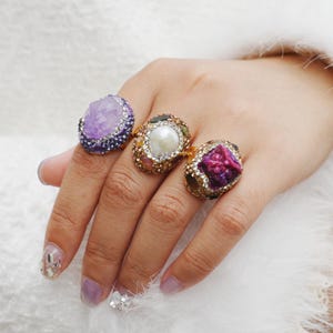 Può includere: Tre anelli ornati con grandi pietre preziose colorate sono esposti su una mano. Gli anelli presentano una grande pietra viola, una perla e una pietra rosso intenso, ciascuna circondata da gemme più piccole e incastonata su fasce d'oro. La mano ha unghie curate.