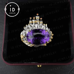 May include: A silver and gold ring featuring a large, oval-cut purple gemstone. The ring is designed with a castle motif, with small towers and gold accents. The ring is presented in a black jewelry box.