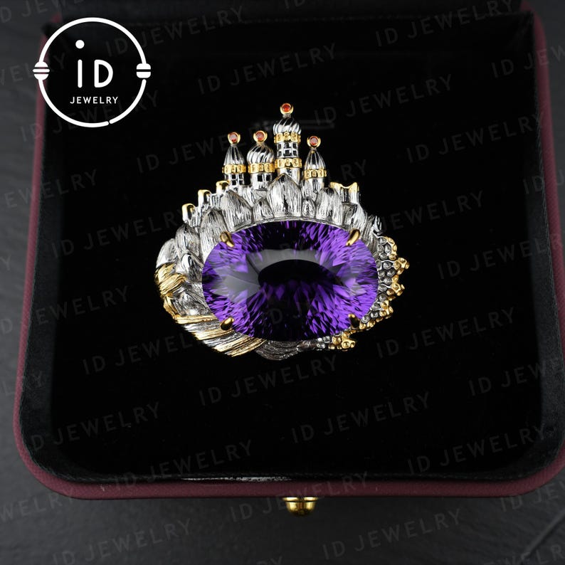 May include: A silver and gold ring featuring a large, oval-cut purple gemstone. The ring is designed with a castle motif, with small towers and gold accents. The ring is presented in a black jewelry box.