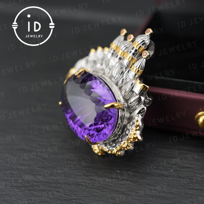 May include: A silver and gold pendant with a large, oval-cut purple gemstone. The pendant has intricate detailing, including a crown-like design with small red accents. The ID Jewelry logo is visible in the upper left corner.