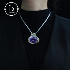 May include: A silver necklace with a large, oval, purple gemstone pendant. The pendant is framed with gold and silver accents, and has a castle-like design. The necklace is worn against a black background, with the "id JEWELRY" logo in the upper left corner.