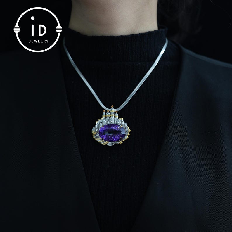 May include: A silver necklace with a large, oval, purple gemstone pendant. The pendant is framed with gold and silver accents, and has a castle-like design. The necklace is worn against a black background, with the "id JEWELRY" logo in the upper left corner.