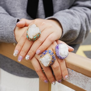 Puede incluir: Se muestran tres anillos grandes y ornamentados con centros de cristal blanco. Uno tiene detalles turquesa, otro azul y dorado, y el tercero morado. Los anillos están en los dedos de una persona, mostrando las joyas.