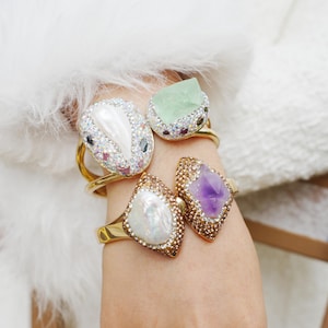 Può includere: Quattro braccialetti a polsino dorati, ognuno ornato da una gemma unica. Uno presenta una pietra bianca iridescente, un altro un cristallo verde chiaro, e altri due mostrano una perla bianca e un'ametista viola. Le gemme sono circondate da piccoli cristalli scintillanti.